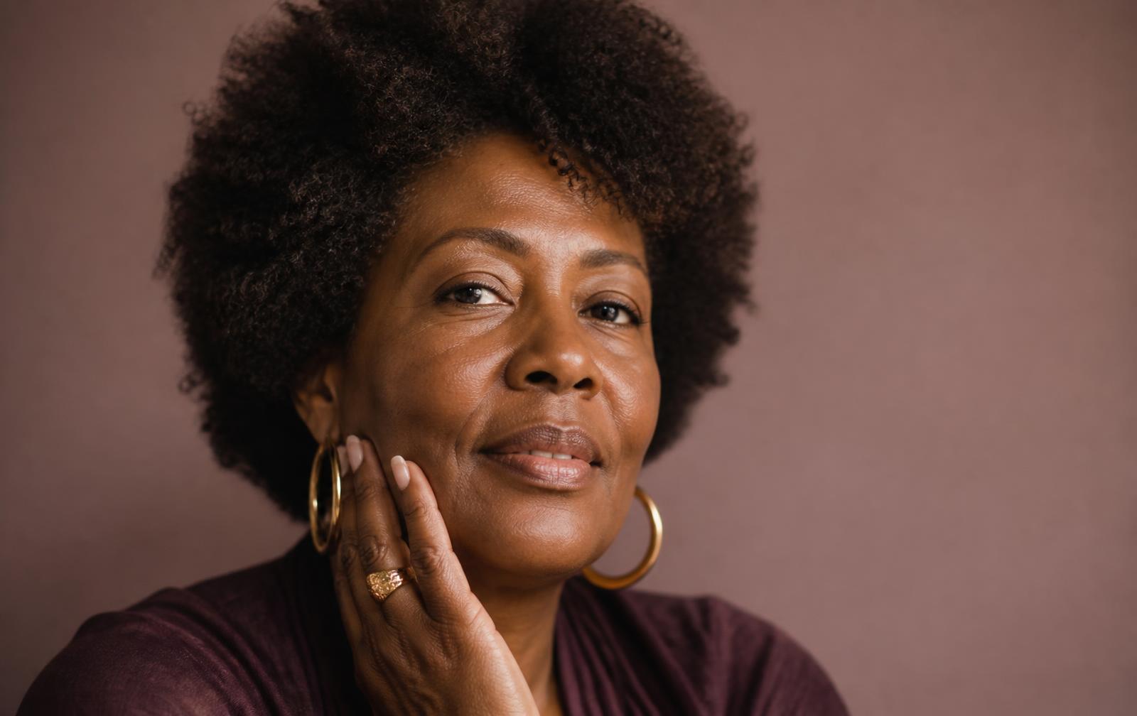 A Black woman in her late forties with luminous skin, resting a hand on her cheek in soft natural light